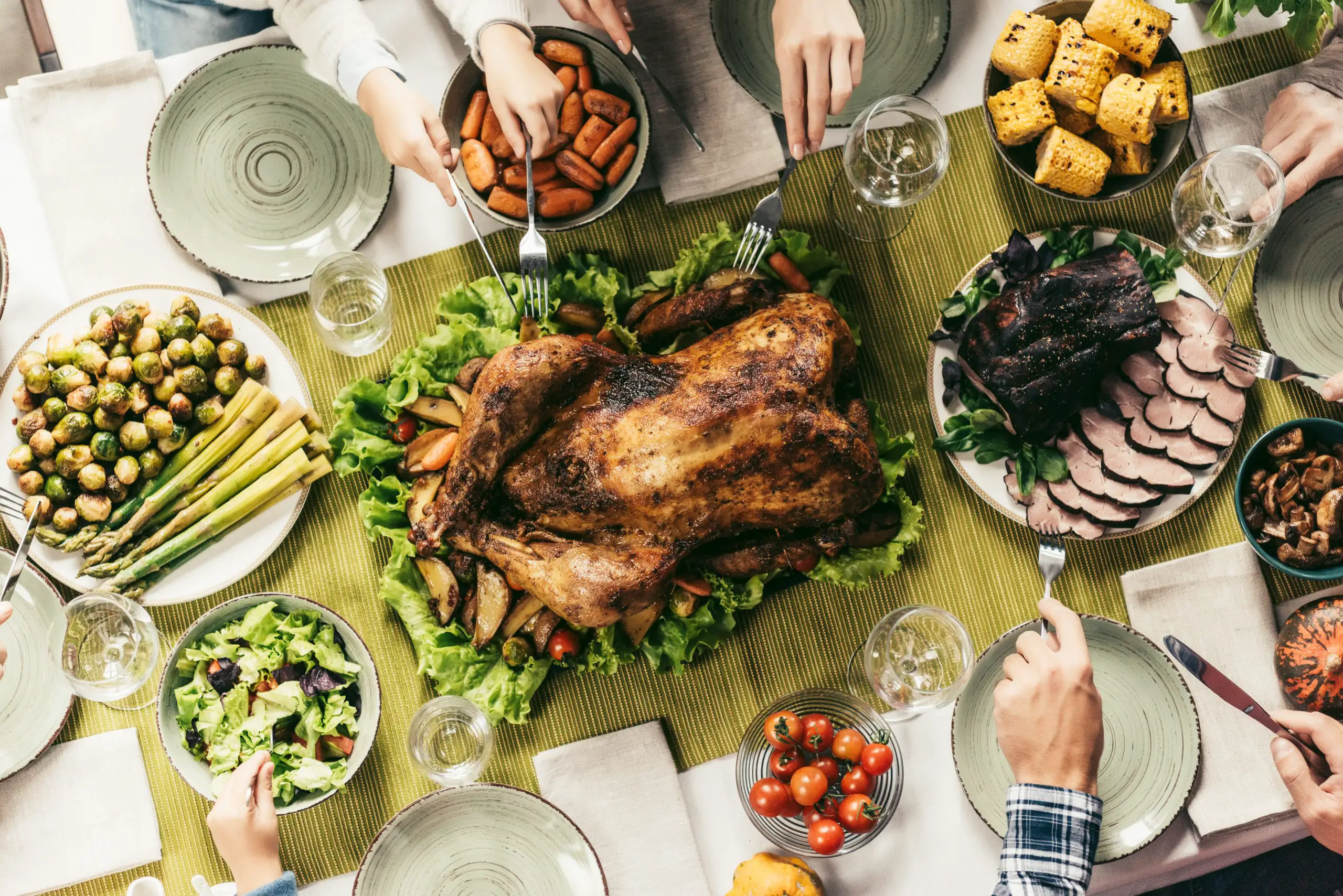 a thanksgiving dinner spread, photo taken from above the table. There is a whole turker, a platter of meat, corn on the cob, green salad, and aspragus and brussels sprouts. People sitting off to the side are reaching in with forks.