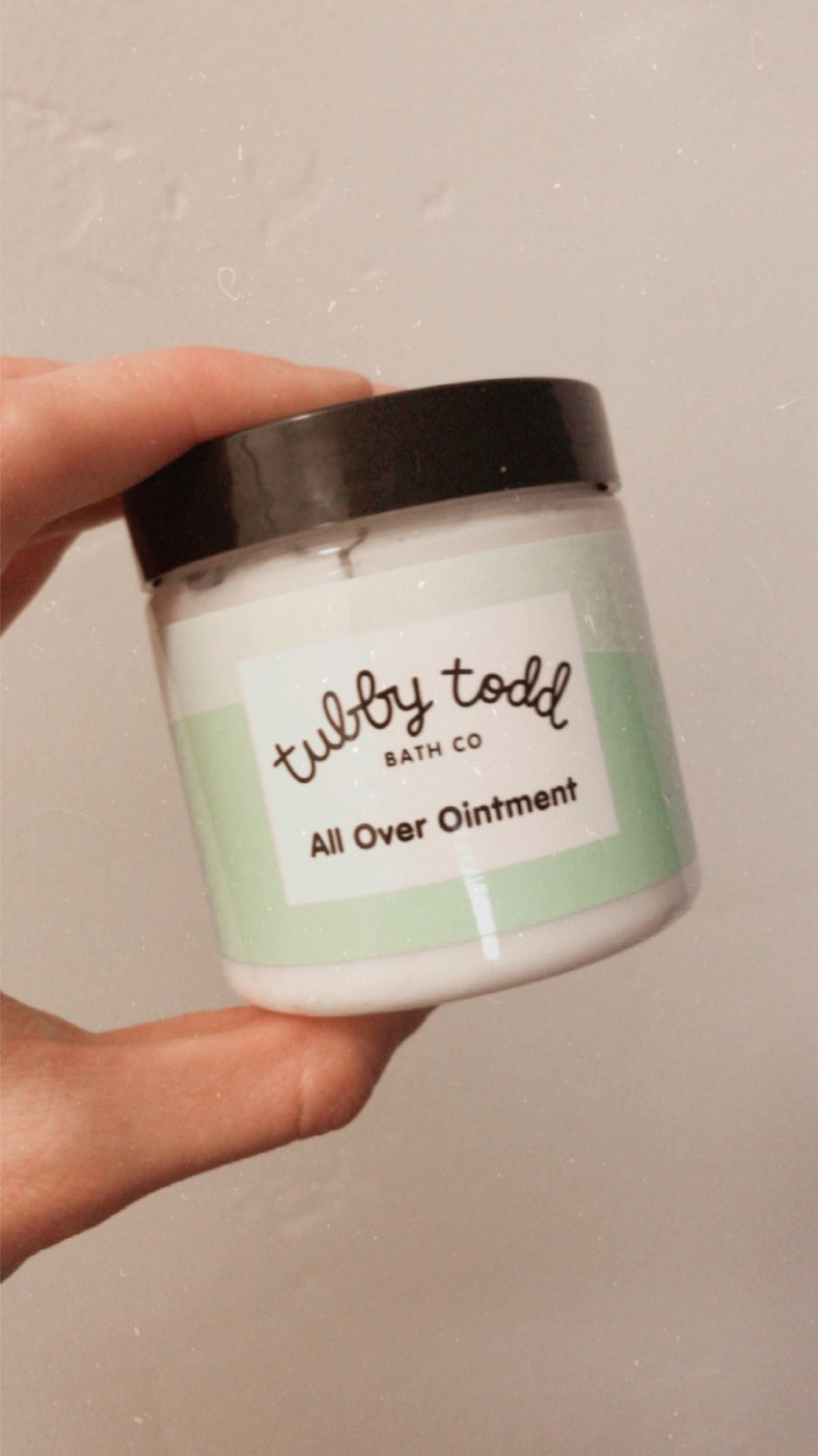 a hand holding a container of tubby todd all over ointment against a white wall background