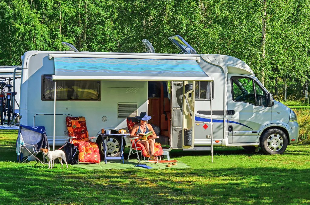 a midsize RV set up at a campground. A woman sits outside with her dog, reading a book