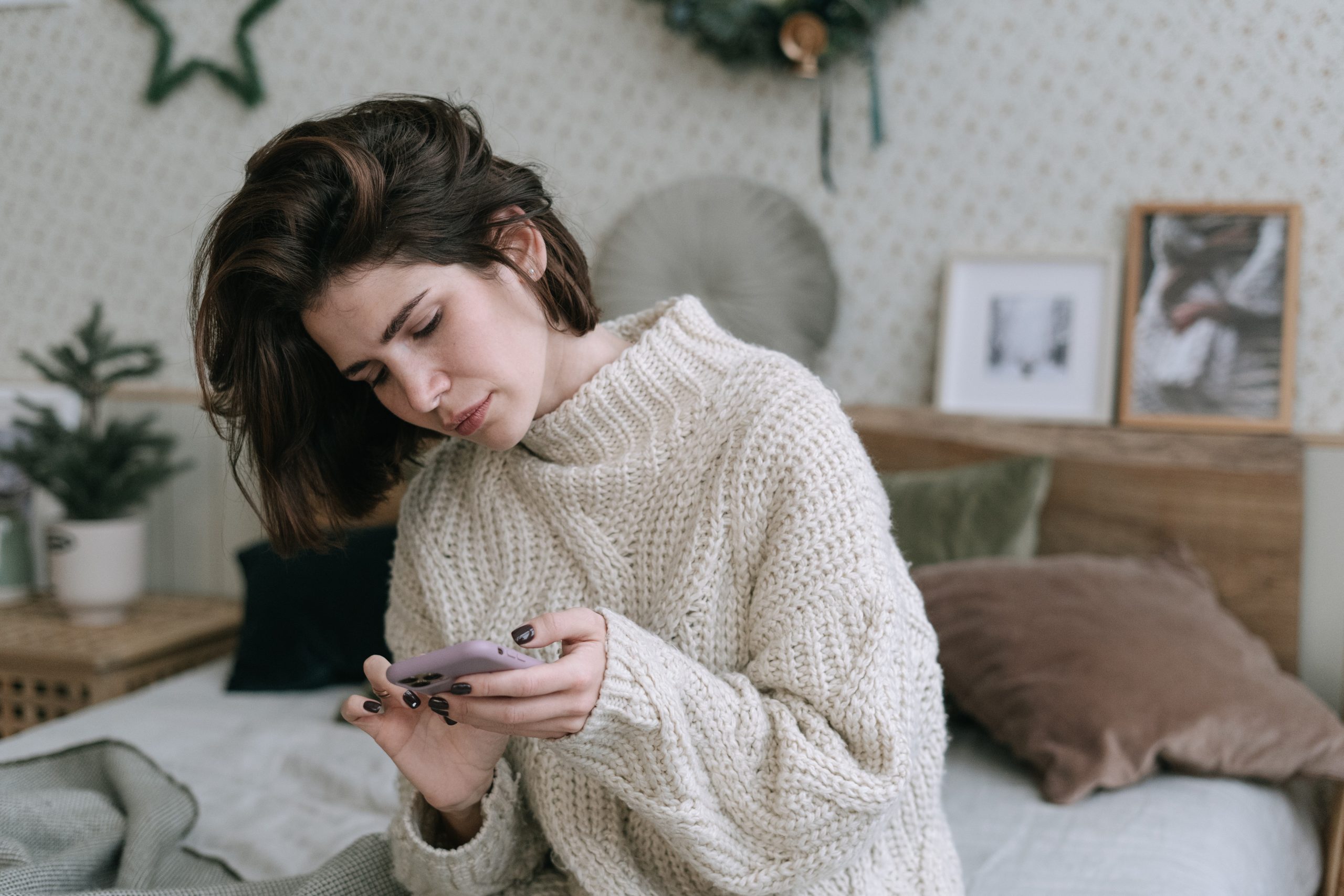 woman in chunky white sweater sitting on a bed, using her phone