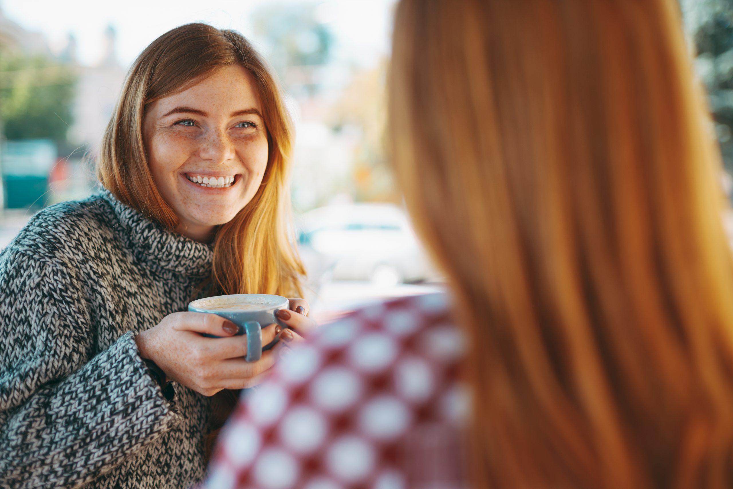 a smiling young woman drinking coffee and speaking to another young woman whos face we cannot see
