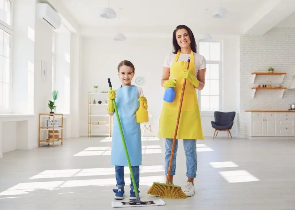 A mother and daughter are holding cleaning products, mops, and brooms.  They're ready to start doing their chores!