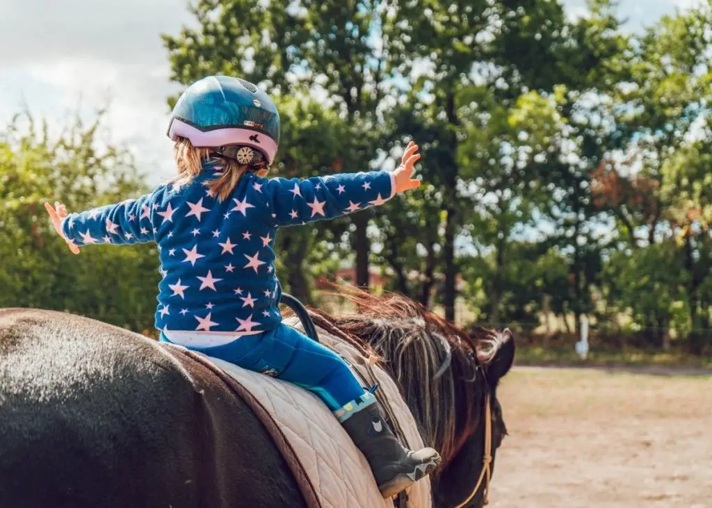 A toddler girl wearing a jacket with stars on it has her arms outstretched as she's riding her horse.  She's wearing a colorful and fun children's horse riding helmet.