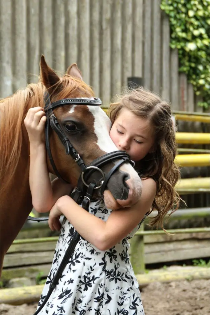 A young girl with brown curly hair is snuggling her horse.