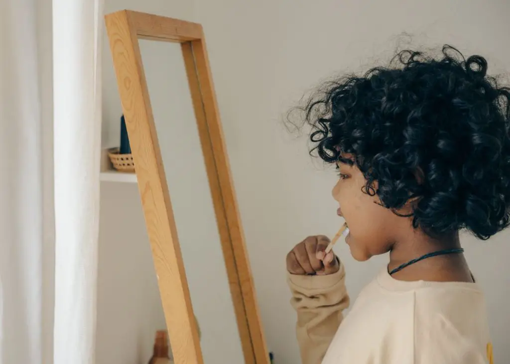 A curly haired little boy is looking in a mirror brushing his teeth.