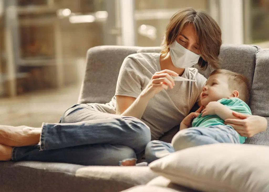 A mom wearing a mask is looking at a thermometer while sitting on a couch with her arm around her young son.
