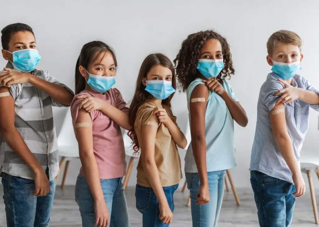 Five kids are holding up their sleeves showing a bandaid where they got a vaccine.