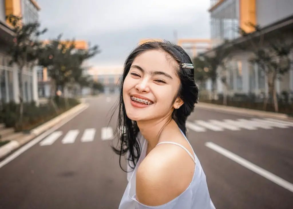Here's a teenage girl with black hair.  She has braces and she's standing in the middle of a city street smiling at the camera.