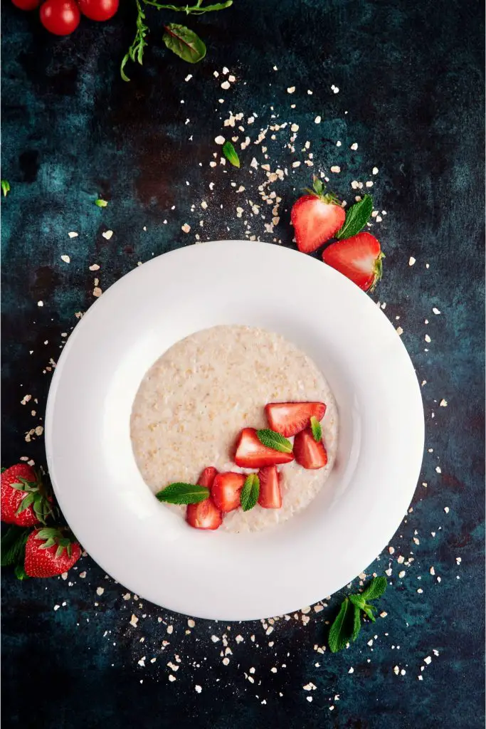 This is an overhead shot of oatmeal with cut strawberries in a white bowl.  Strawberries and oats surround the bowl.  It has a blue background.