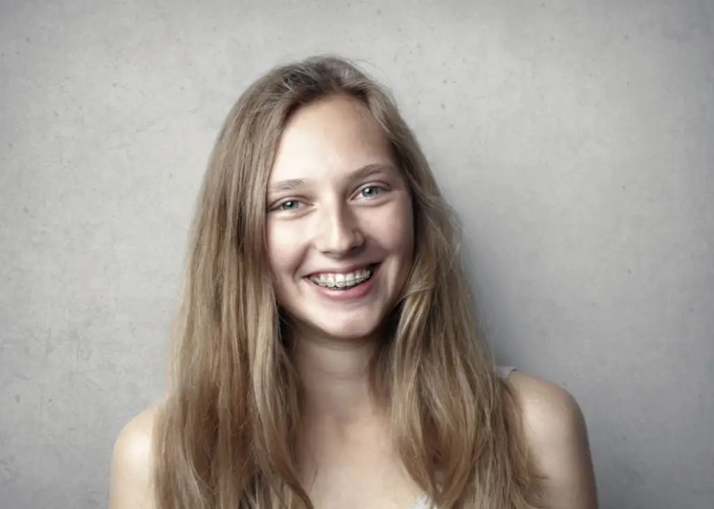 A girl with light brown hair is smiling at the camera.  She's wearing a tank top and she has braces on her teeth.