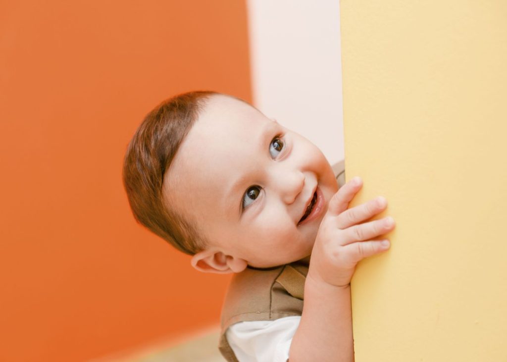 A toddler boy is peering over a yellow wall.