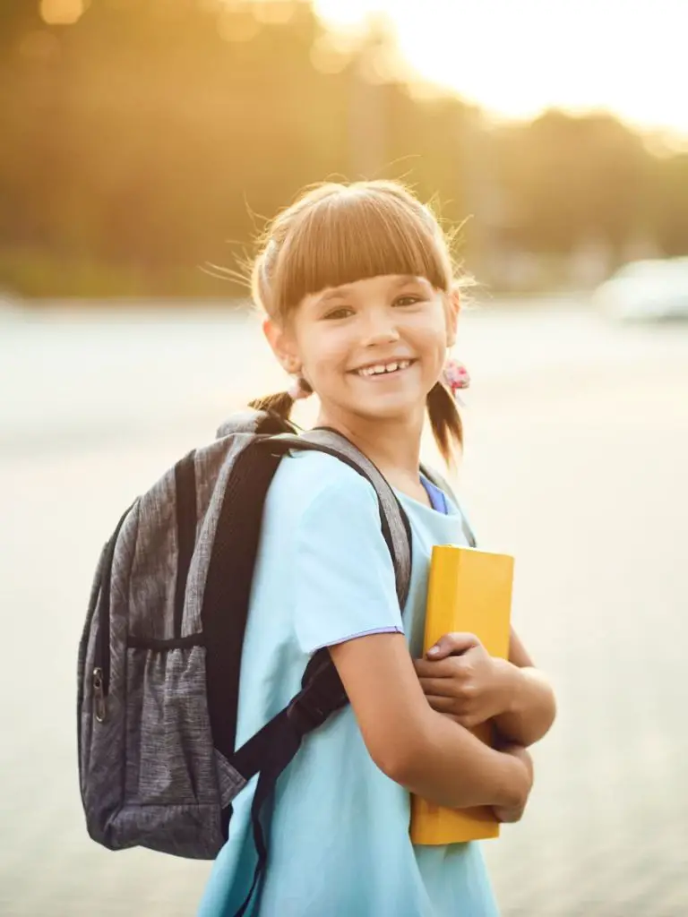 A young girl with short brown hair is wearing a backpack, holding a book, and smiling.